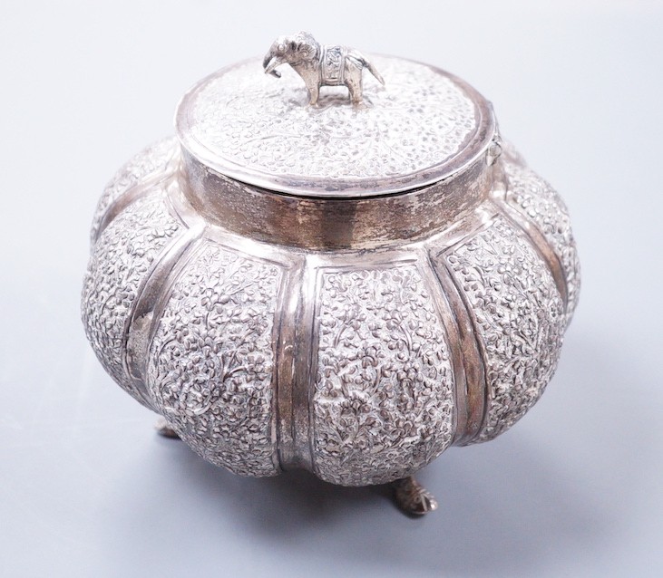 An Indian embossed lobed white metal circular pot and cover, with elephant finial, on three dolphin feet, height 10cm, 197 grams.